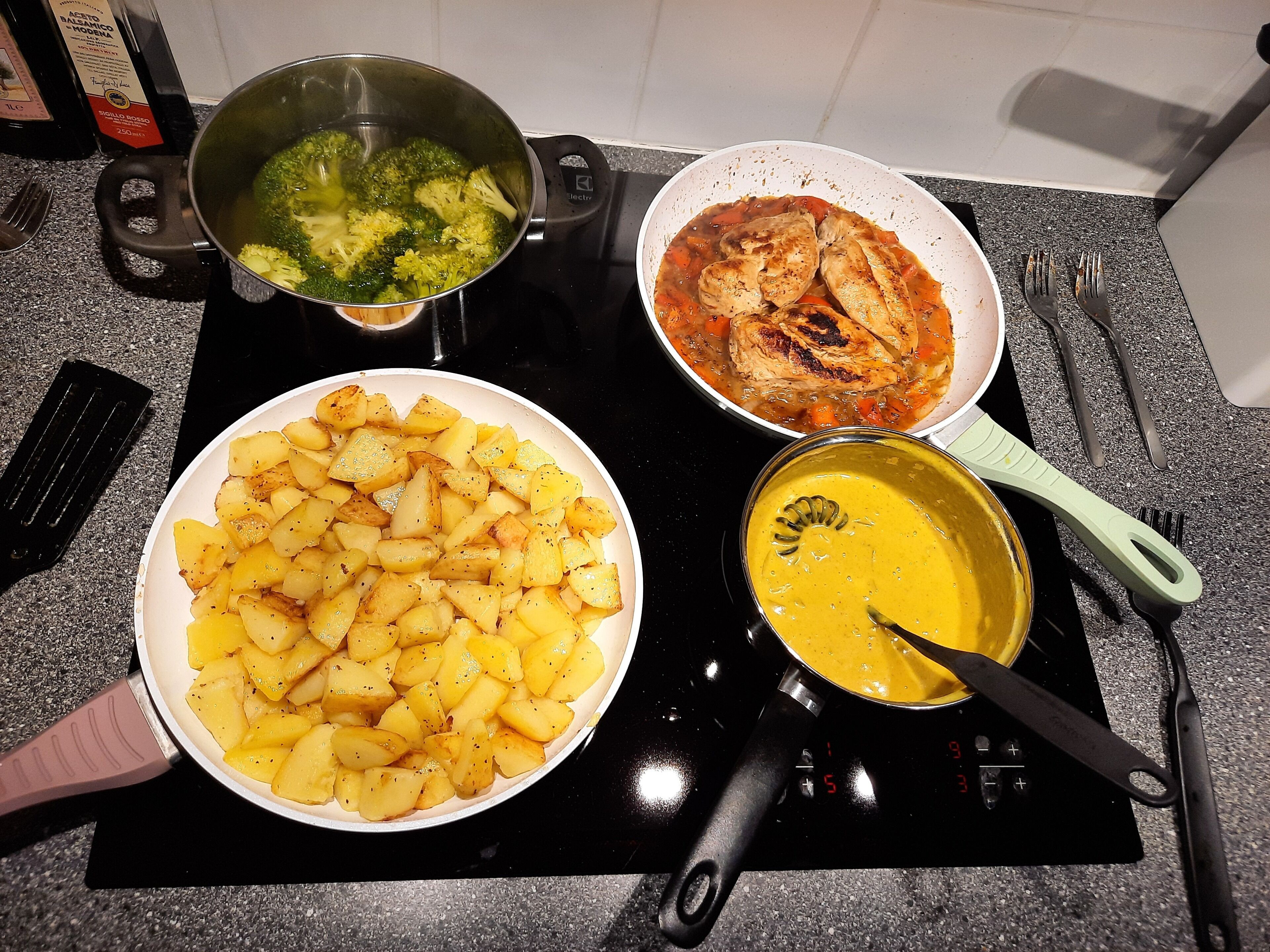Una encimera de cocina con cuatro ollas y sartenes que contienen diversos platos, incluyendo papas, brócoli y una salsa amarilla.
