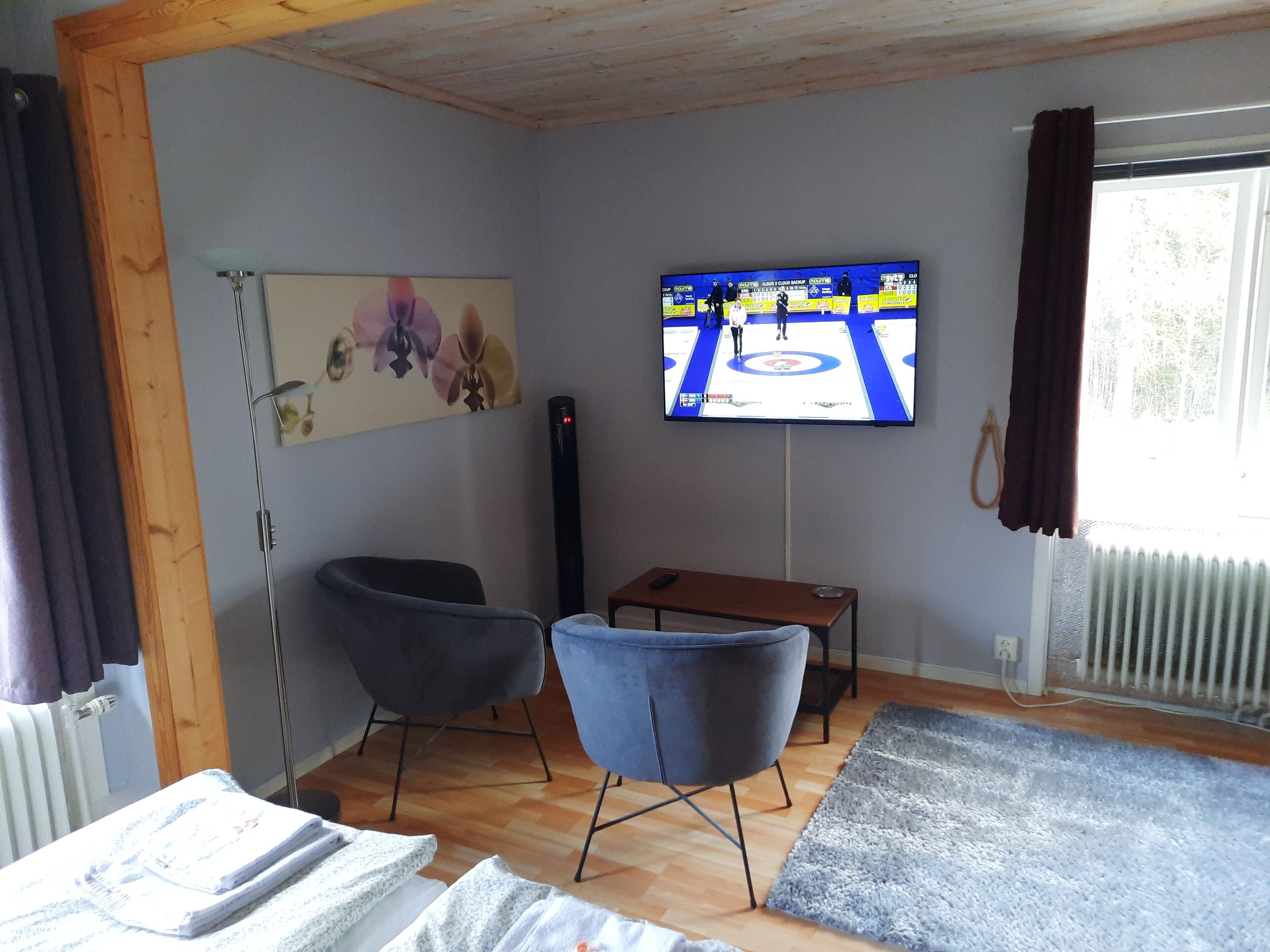 Una sala de estar con un televisor montado en la pared, un sillón azul, un banco de madera y un radiador.