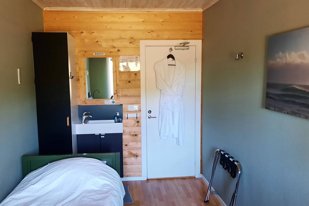 Een slaapkamer met een bed, een houten plafond, een badkamer met een wastafel en spiegel, en een deur waar een handdoek aan hangt.