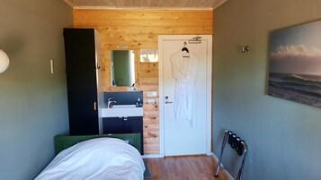 A bedroom with a wooden ceiling, a bed, a bedside table, a mirror, a bathroom with a sink and a towel rack, and a painting of the ocean on the wall.