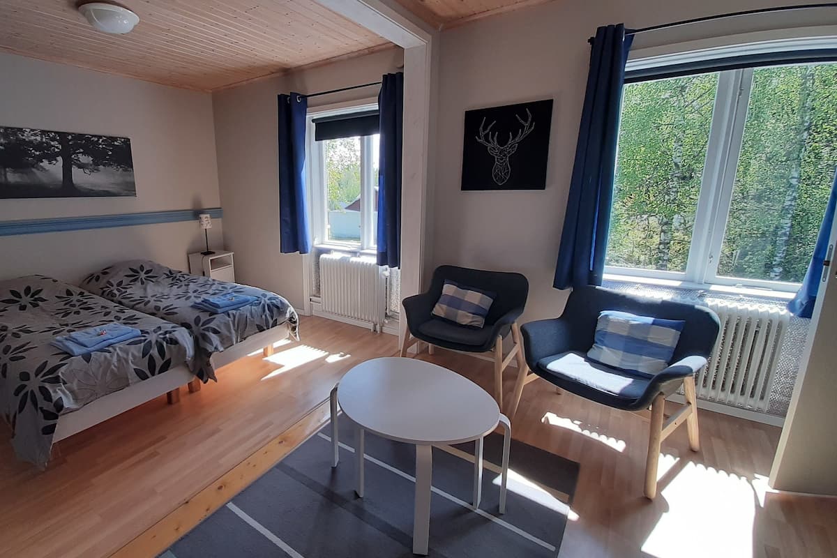 Een kamer met een bed, een stoel, een klein tafeltje, een radiator, een raam met gordijnen en een aan de muur hangend schilderij.