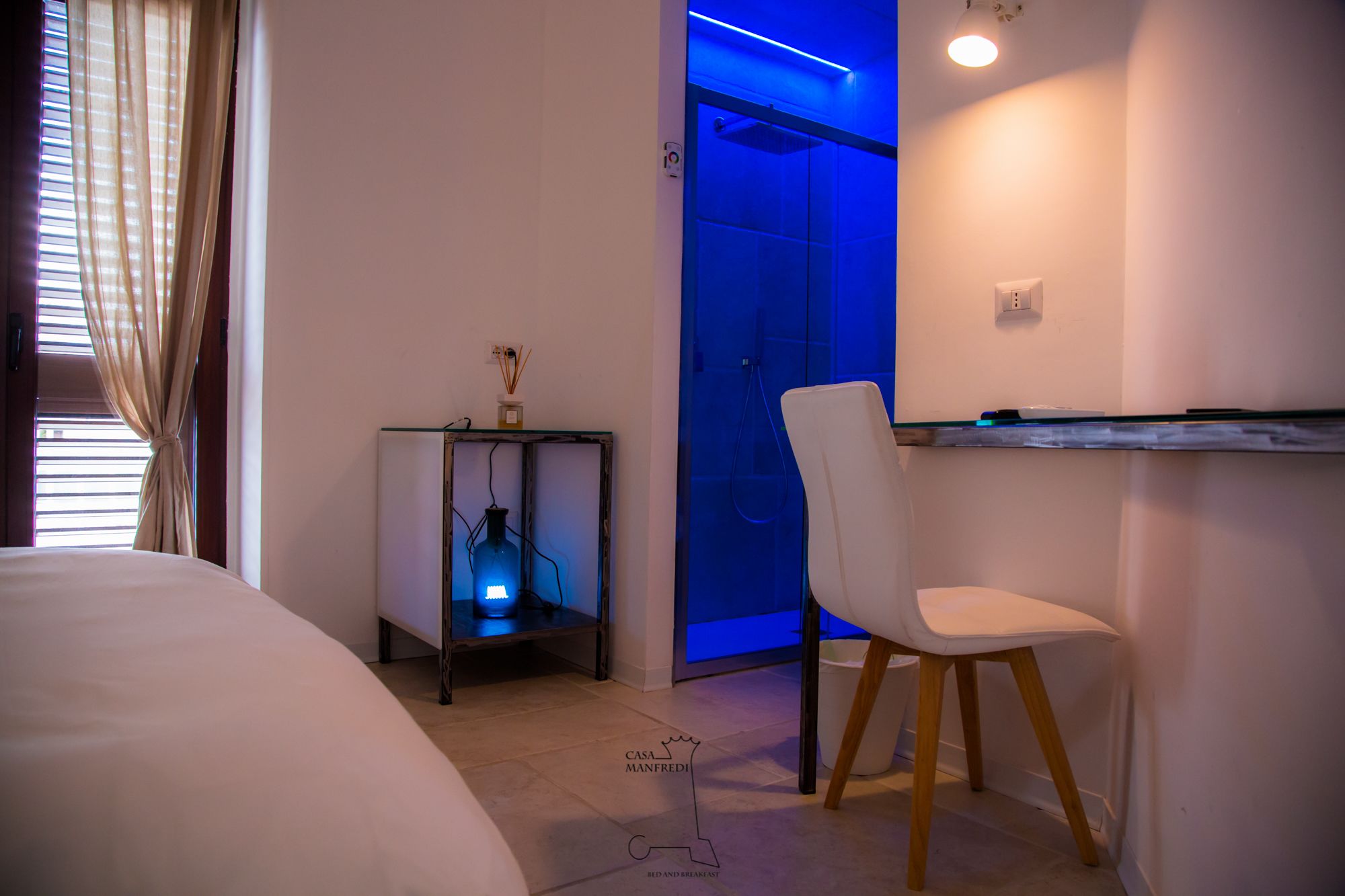 Ein Schlafzimmer mit einem Bett, einem Nachttisch mit Lampe, einem Sessel und einem Badezimmer mit Dusche und blauer Beleuchtung.