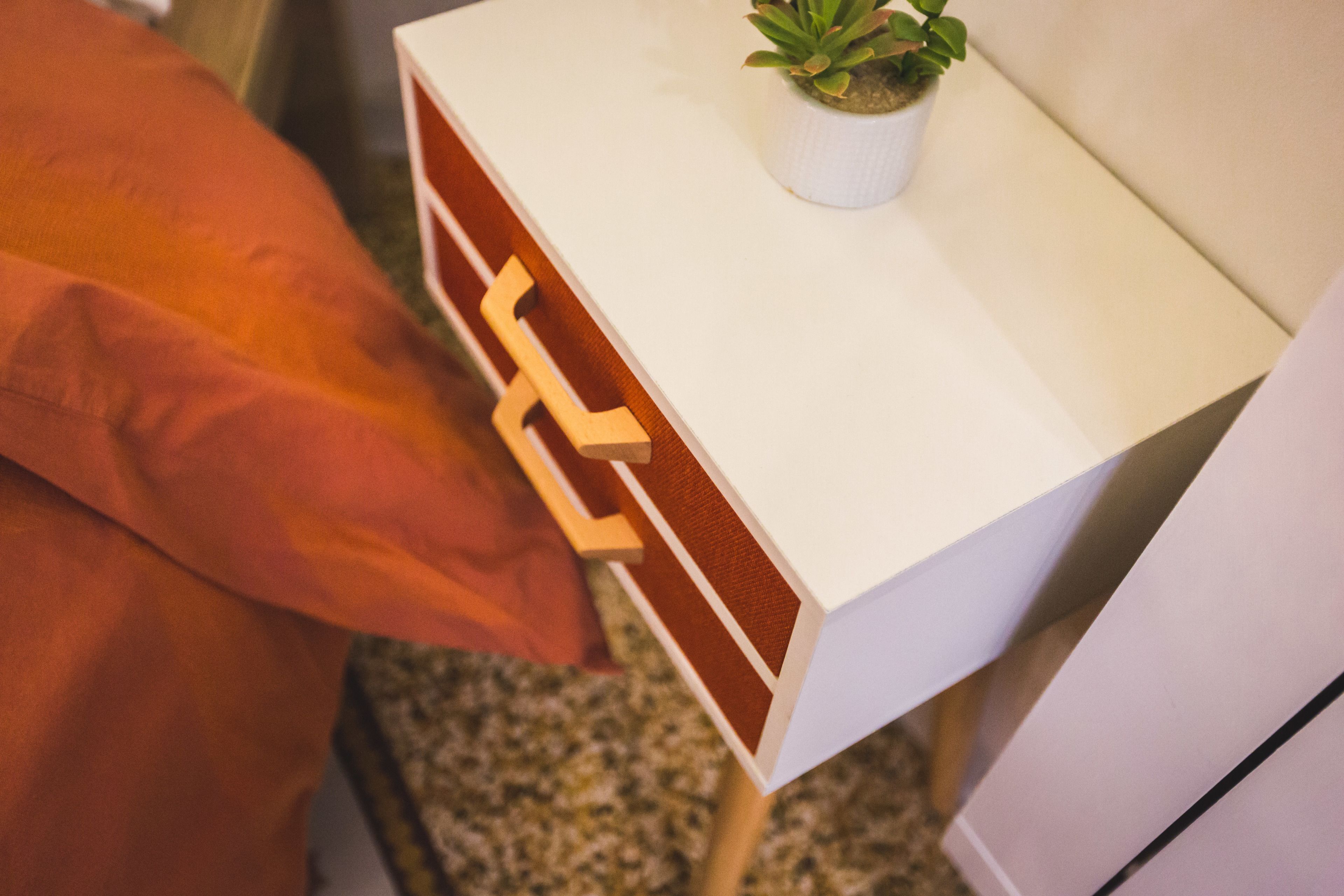 Une table de chevet blanche avec un tiroir, une petite plante en pot et une couverture rouge sur le lit.