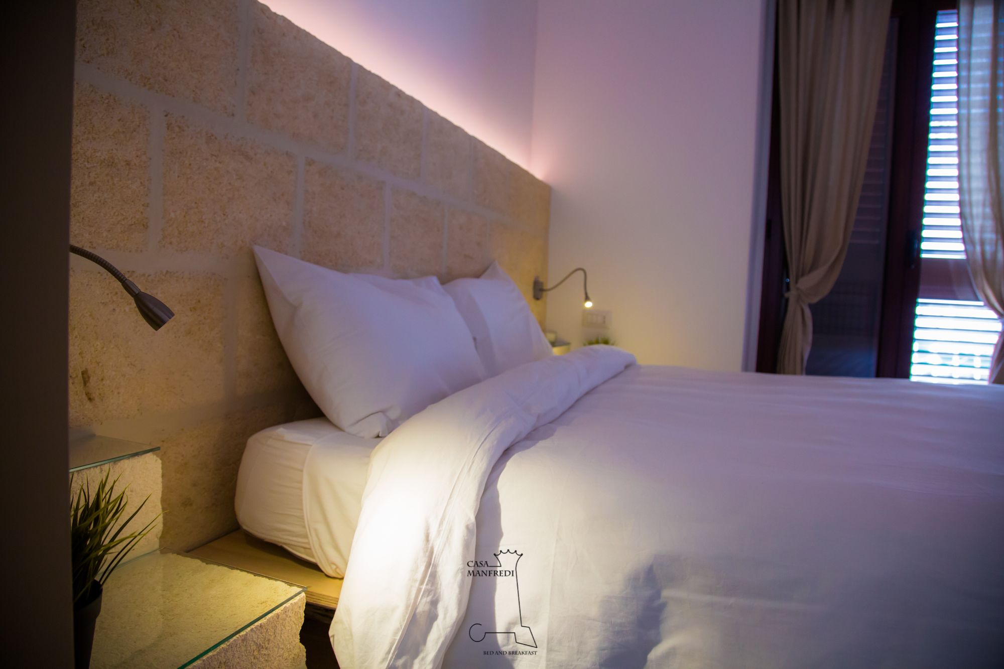 A neatly made bed with white linens, a stone headboard, a bedside lamp, and a small table with a plant.