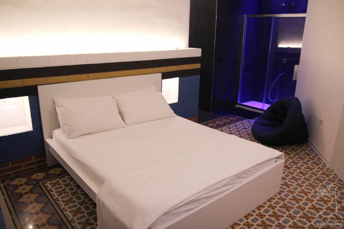 A neatly made bed with white linens in a room with a patterned floor, a dark blue wall, and a shower area with a glass door.