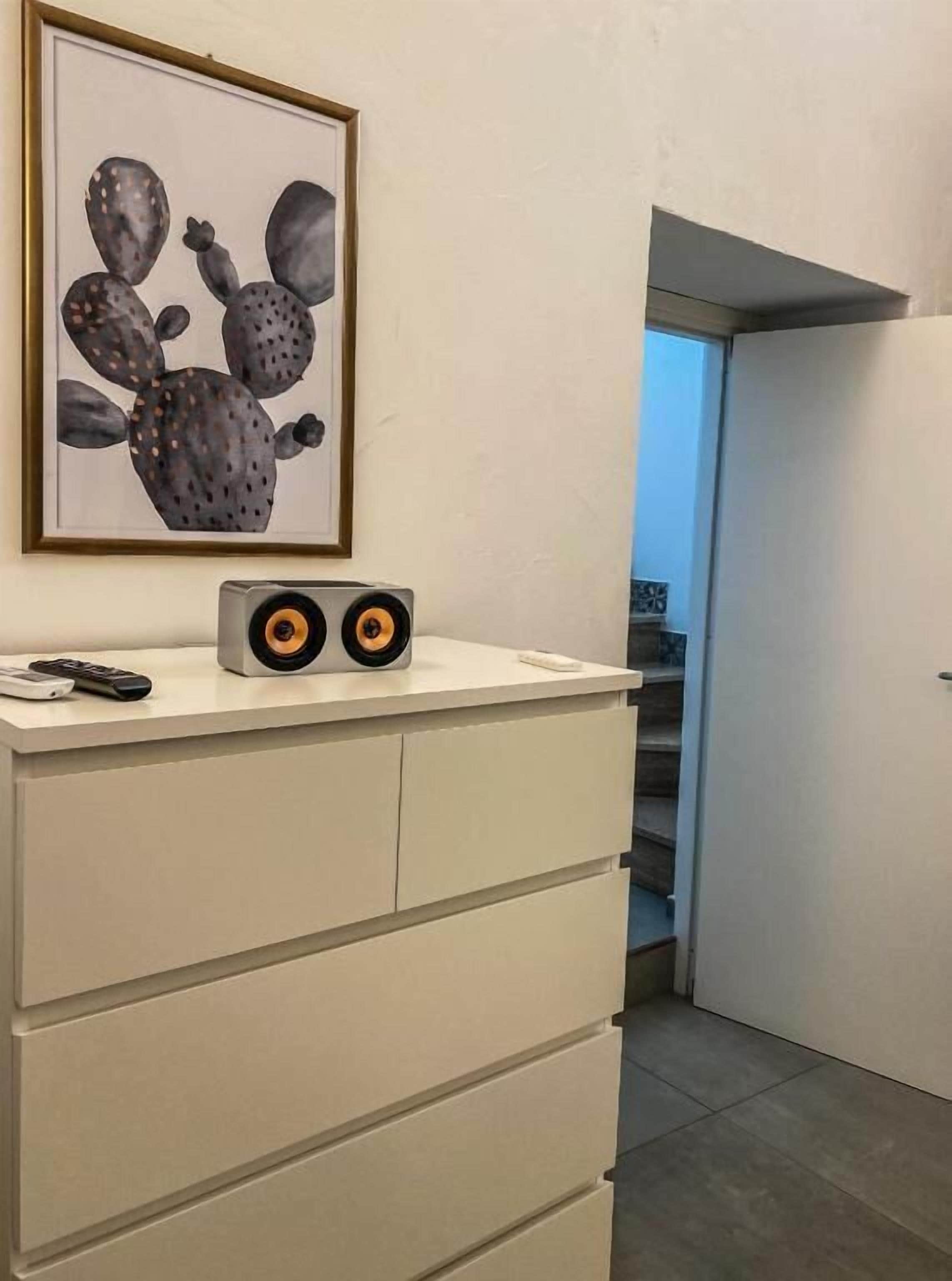 Une commode blanche, au-dessus de laquelle est accrochée une œuvre d’art représentant un cactus encadré, une paire d’enceintes posées sur le dessus et une télécommande à côté des enceintes.