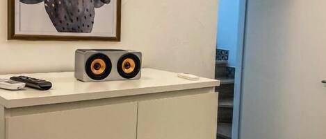 Une commode blanche, au-dessus de laquelle est accrochée une œuvre d’art représentant un cactus encadré, une paire d’enceintes posées sur le dessus et une télécommande à côté des enceintes.