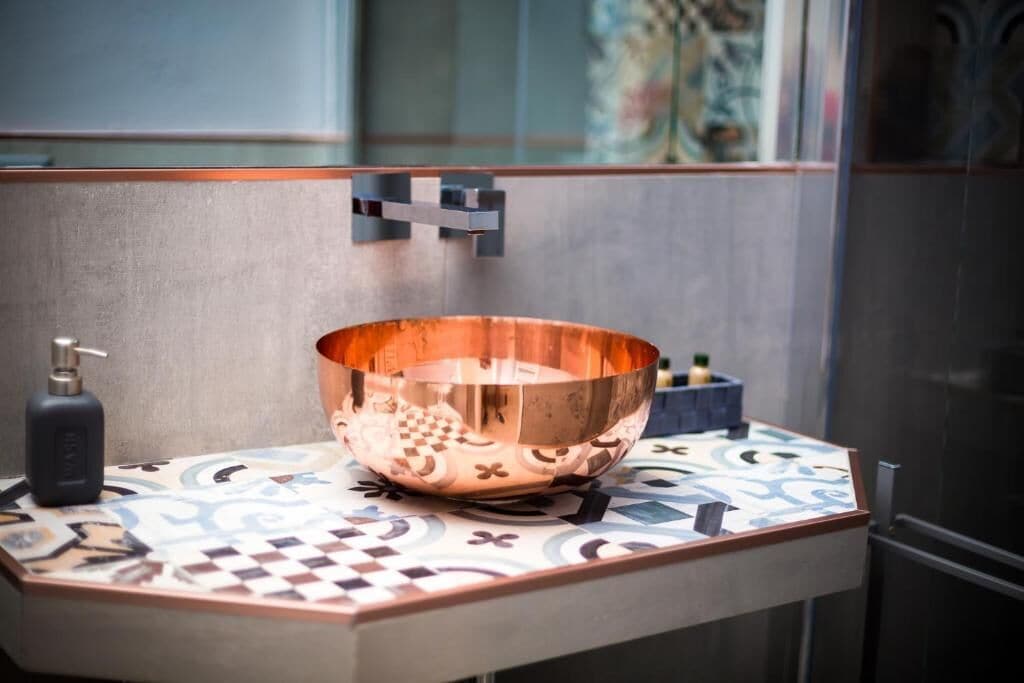 A bathroom with a copper bowl sink, a soap dispenser, and a small blue container on a tiled countertop.