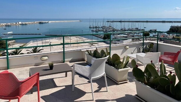 Een terras op het dak met helder uitzicht op zee, voorzien van een zithoek met stoelen en plantenbakken, en een glazen hekwerk.