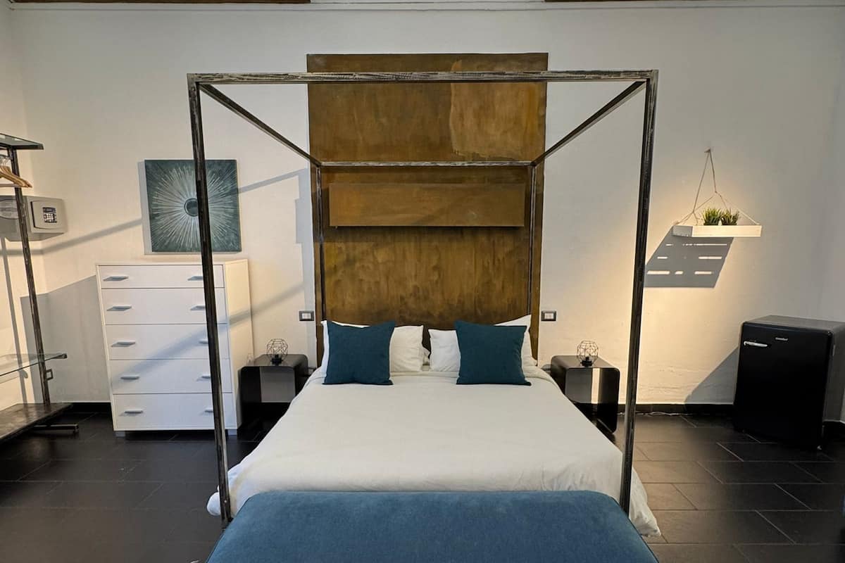 Une chambre à coucher moderne avec un lit à baldaquin, une commode blanche, une table de chevet, un petit bureau et une plante suspendue.