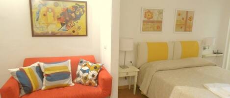 A hotel room with a red sofa, a bed with yellow and white bedding, and two wall-mounted lamps.