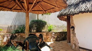 An outdoor patio with a wooden table and chairs, a thatched roof, and a stone wall with plants.