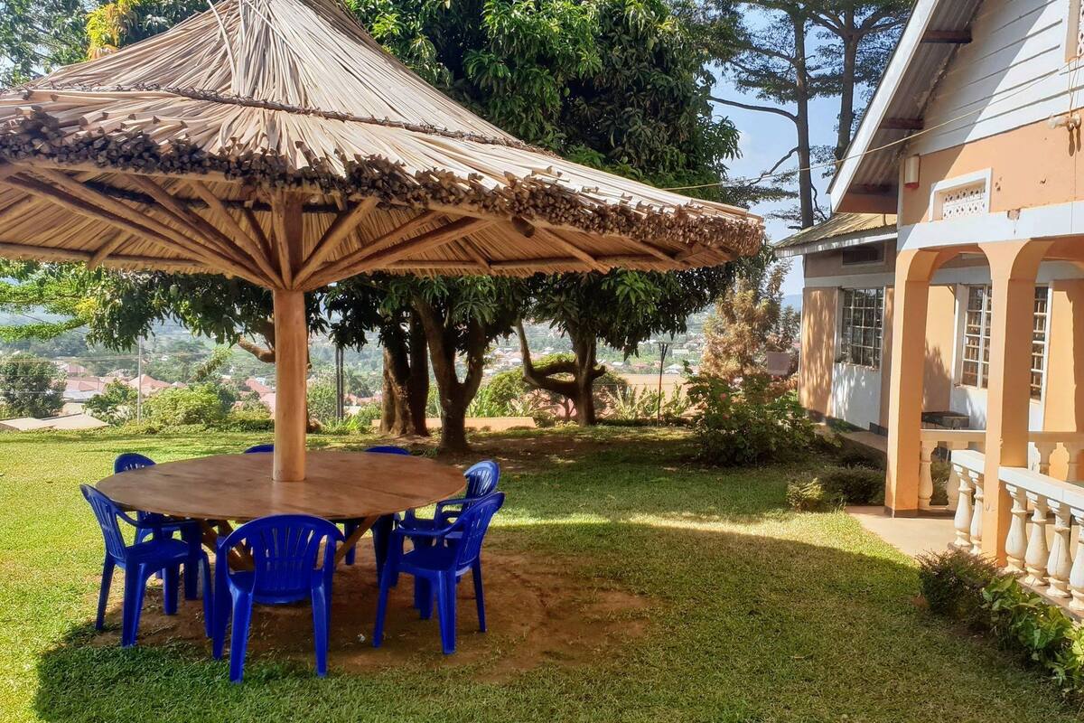 Un área de estar al aire libre con techo, una mesa y sillas de plástico azules, rodeada de césped cuidado y árboles.