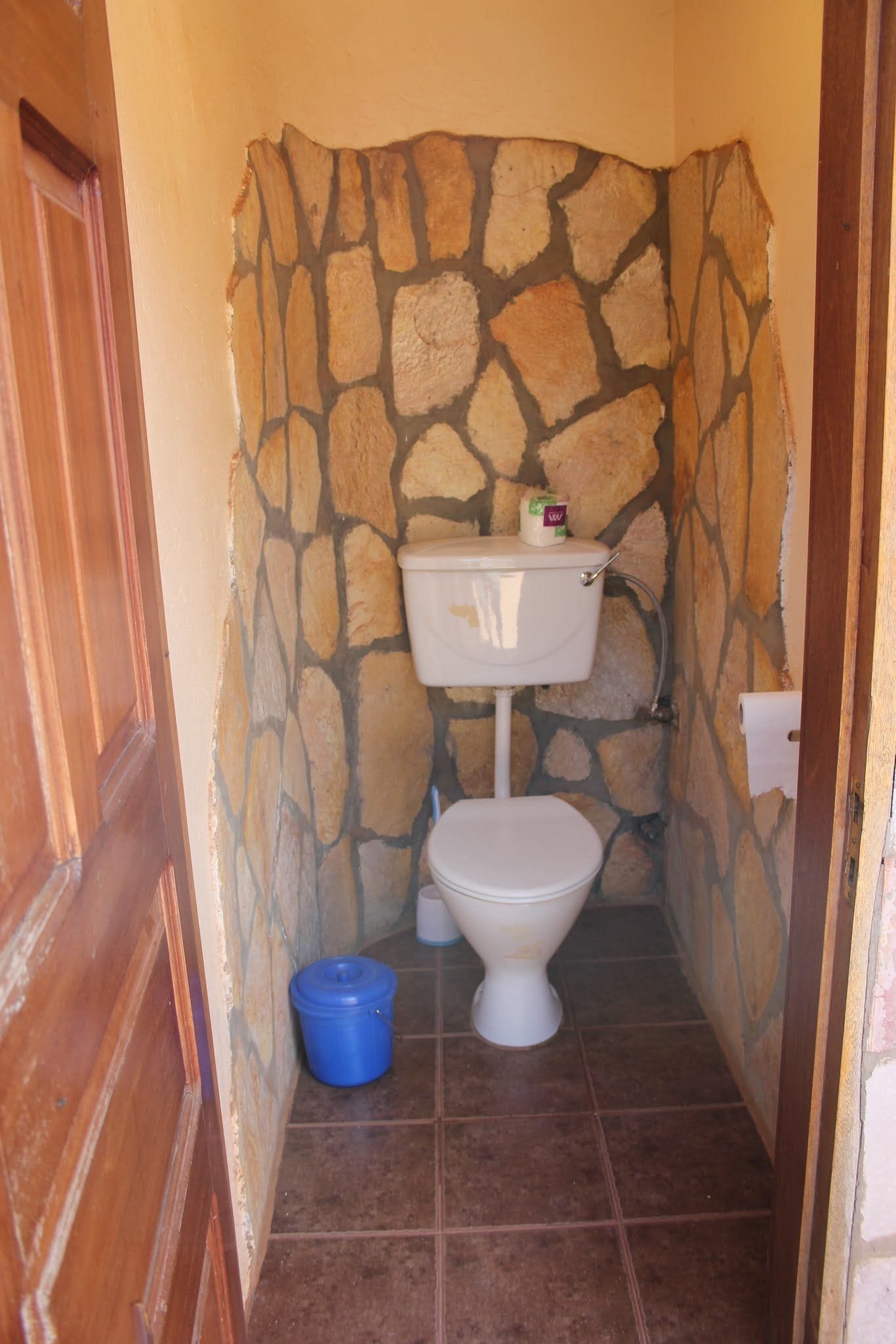 A small bathroom with a stone wall, a white toilet, a blue bucket, and a wooden door.