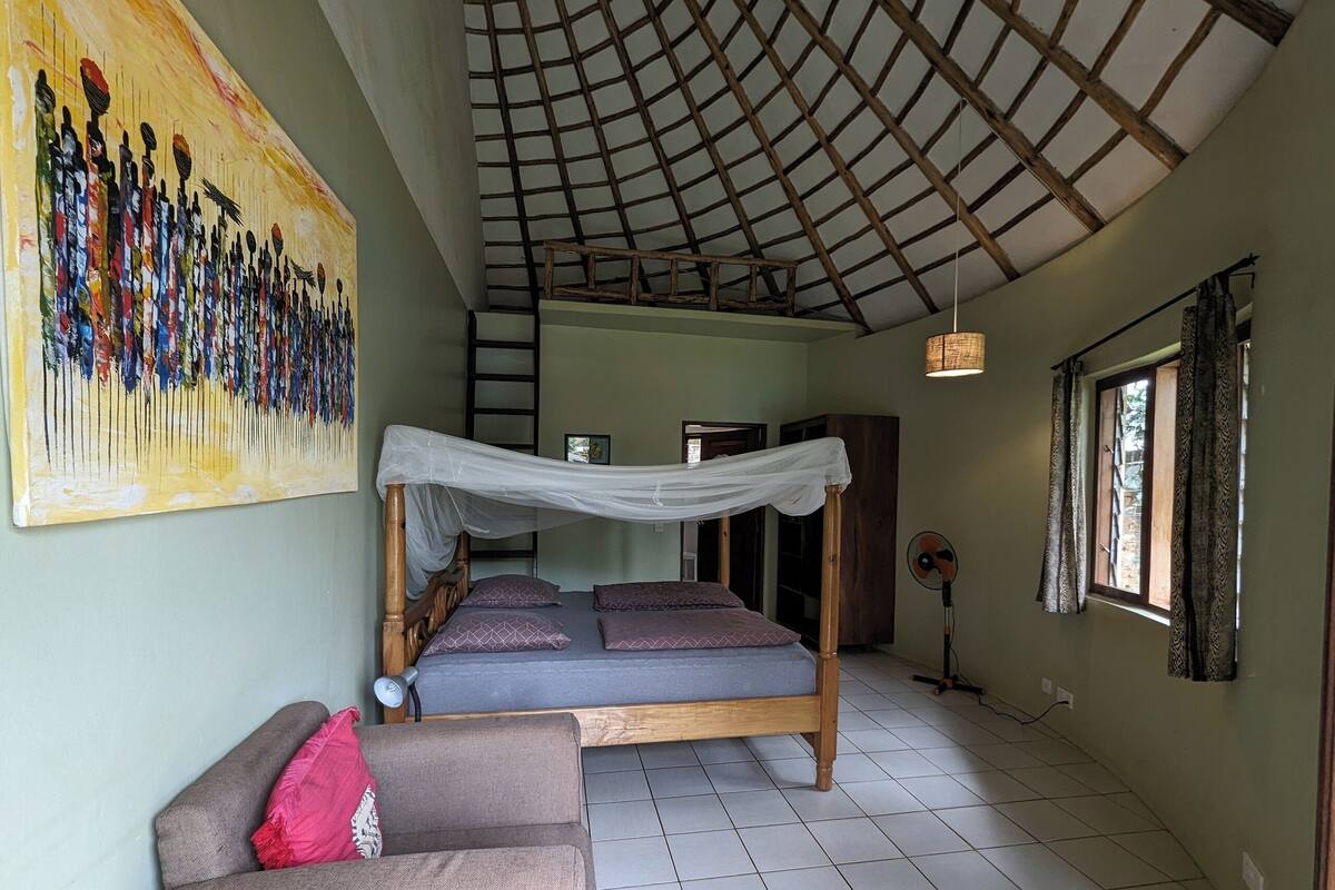 Una habitación con cama, sofá, un cuadro, una escalera, un ventilador y una ventana con cortinas.