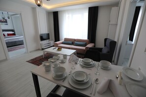A modern hotel room with a dining area set for a meal, a sofa, and a TV in the background.