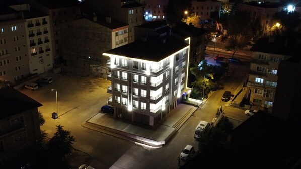 A multi-story building with illuminated windows and a flat roof, surrounded by other buildings and a street.