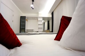 A modern hotel room with a large bed, white bedding, and red pillows. There is a wardrobe, a mirror, and a small table in the room.