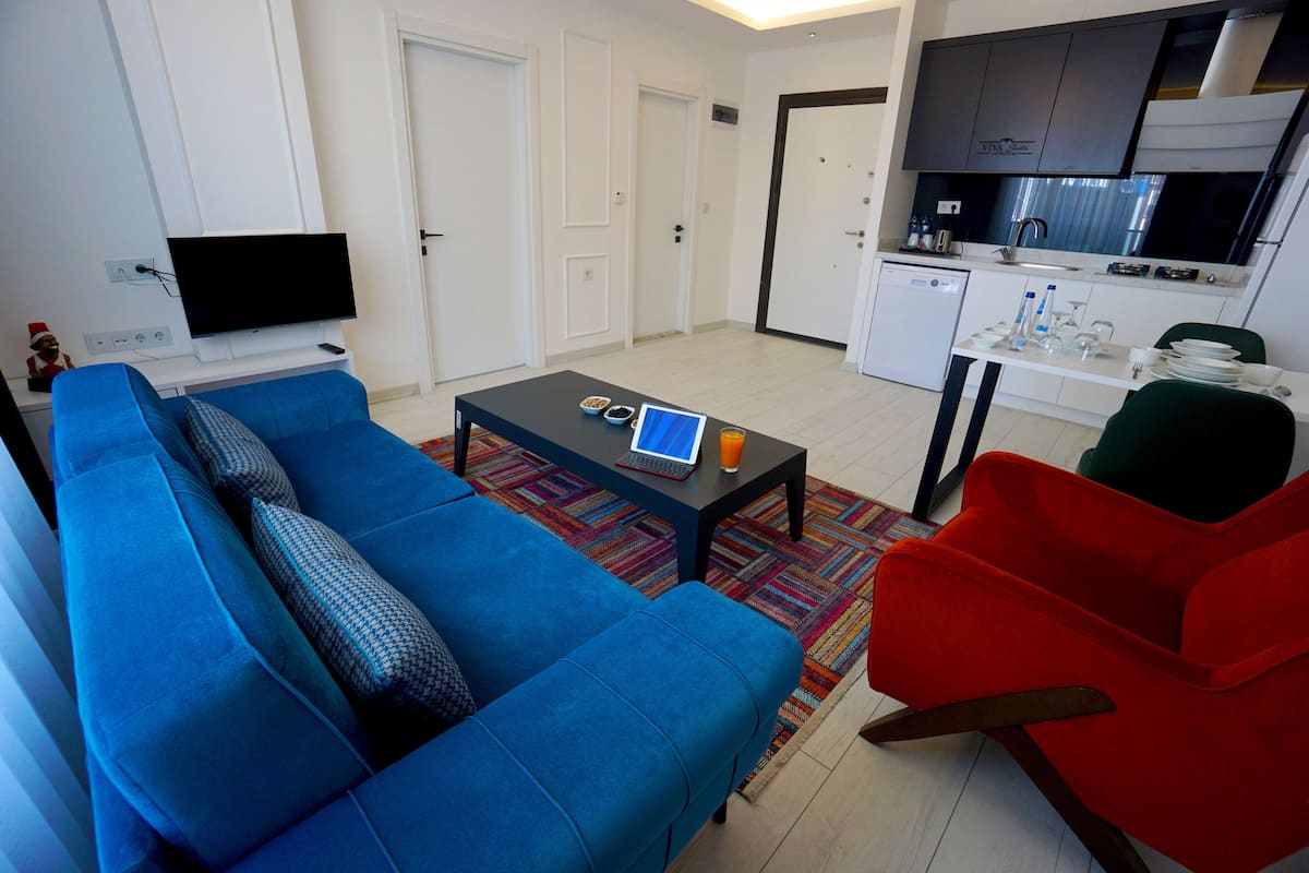 Ein modernes Wohnzimmer mit einem blauen Sofa, einem kleinen Couchtisch und einem roten Sessel. Im Hintergrund ist eine Küchenzeile mit Schränken und einem Spülbecken zu sehen.