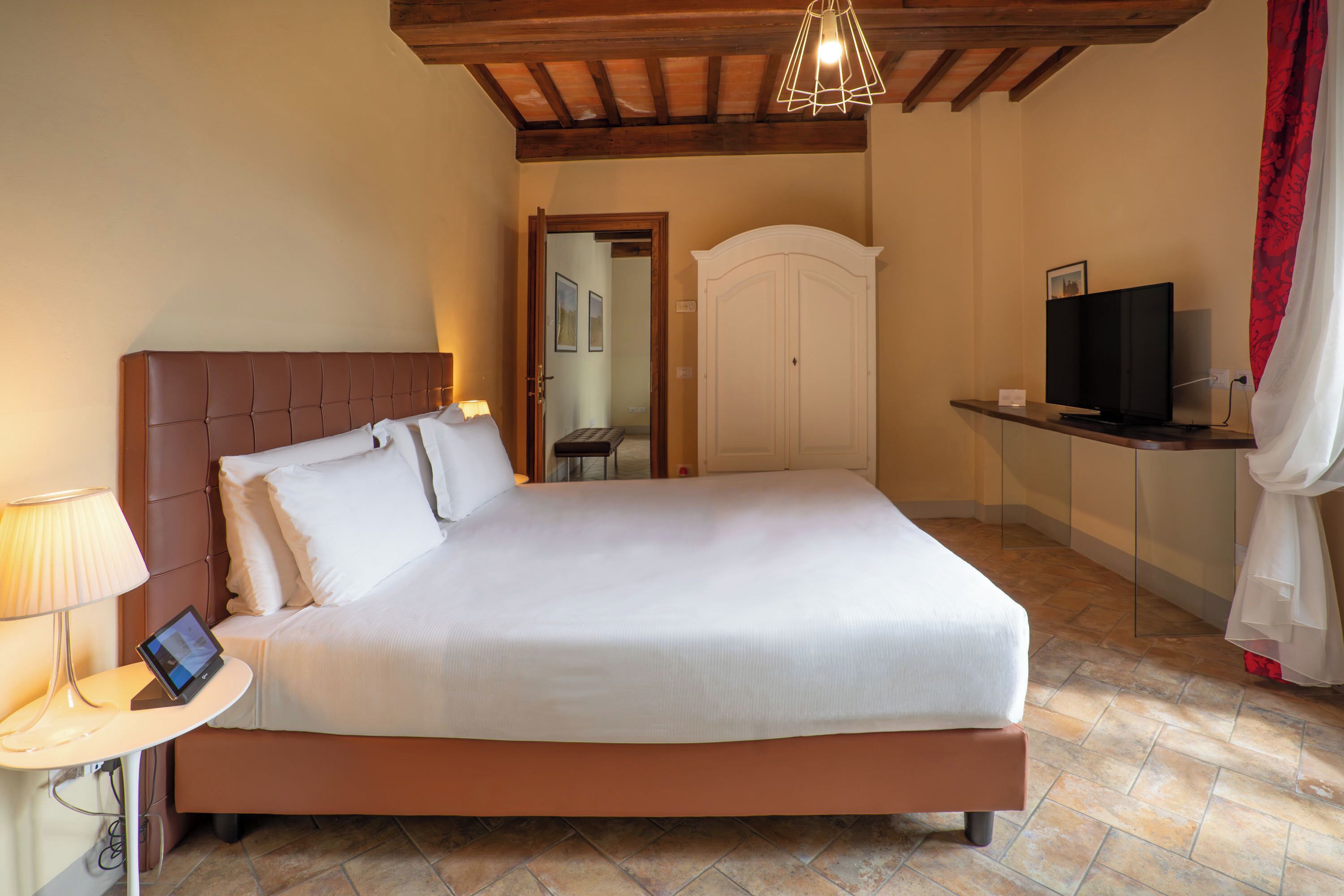 A neatly made bed with a headboard, a bedside table with a lamp and a tablet, a television on a stand, and a wardrobe in a room with tiled flooring and wooden beams on the ceiling.