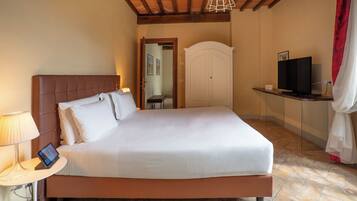A neatly made bed with a headboard, a bedside table with a lamp and a tablet, a television on a stand, and a wardrobe in a room with tiled flooring and wooden beams on the ceiling.
