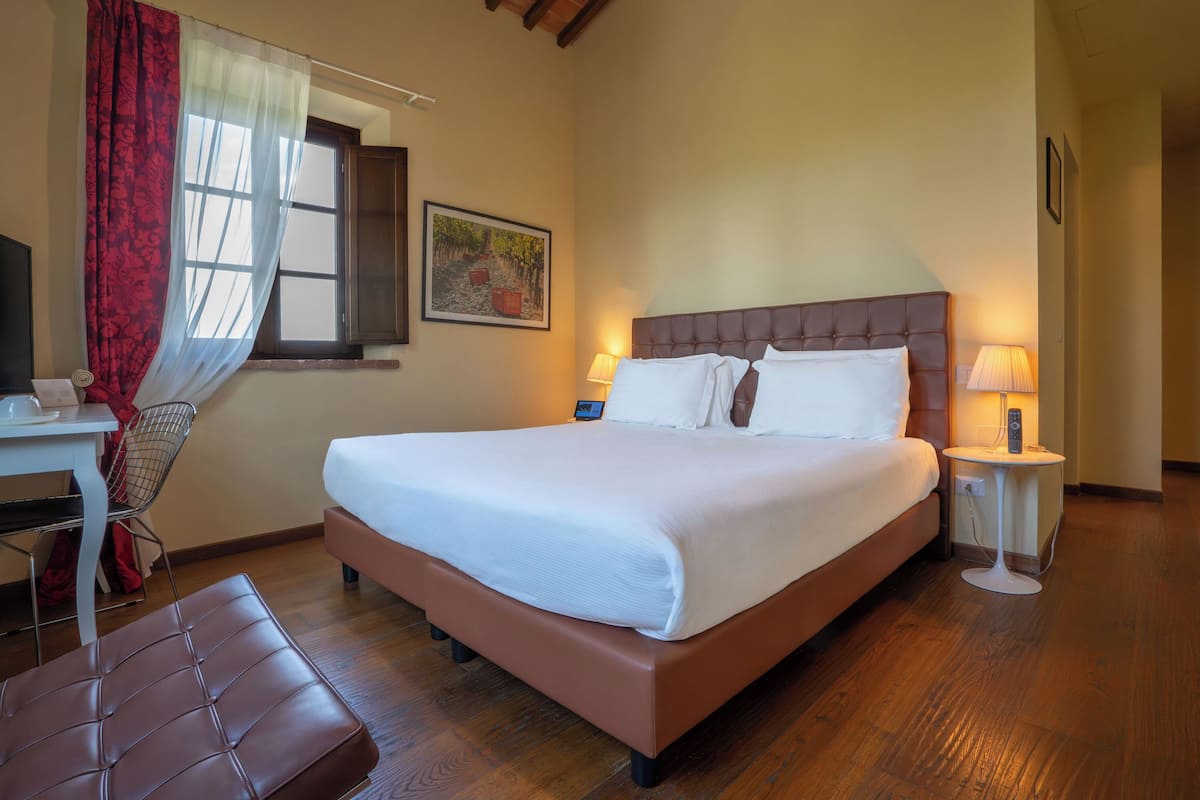 Une chambre d’hôtel comprenant un lit, un bureau, une chaise, une table de chevet avec une lampe et une fenêtre avec des rideaux.