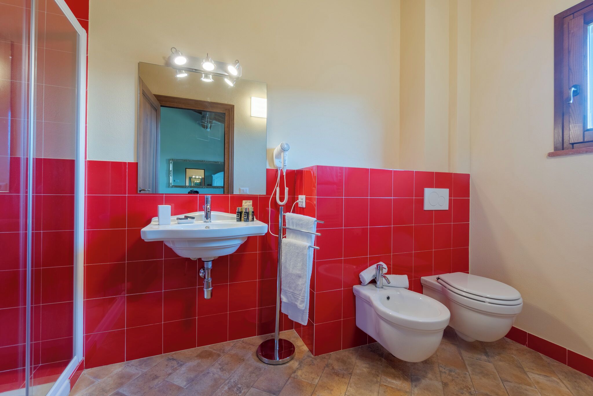 Casa de banho com azulejos vermelhos, lavatório, espelho, porta-toalhas e duas sanitas.