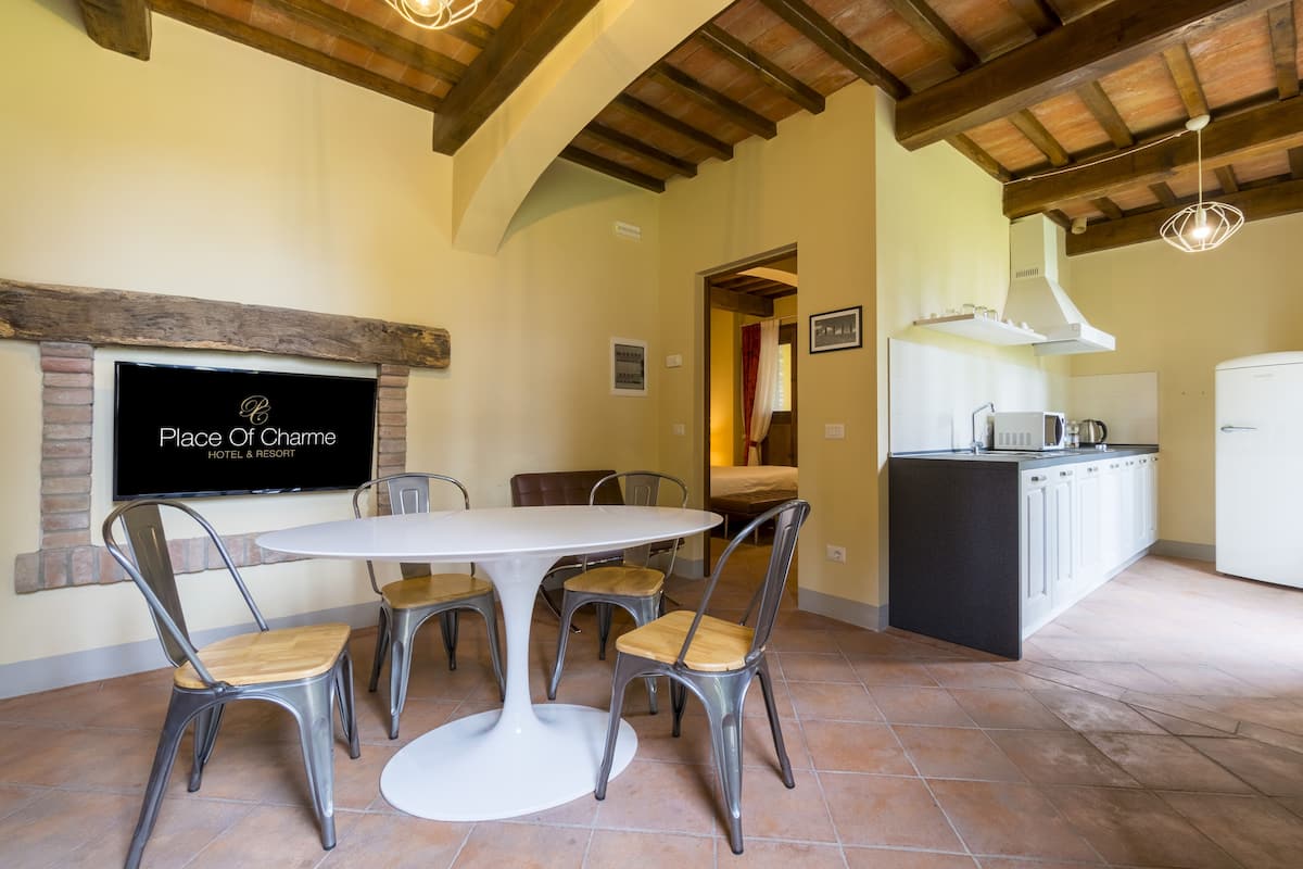 Un coin repas avec une table ronde et six chaises, un téléviseur fixé au mur, et un espace cuisine équipé d’un four à micro-ondes et d’un évier.
