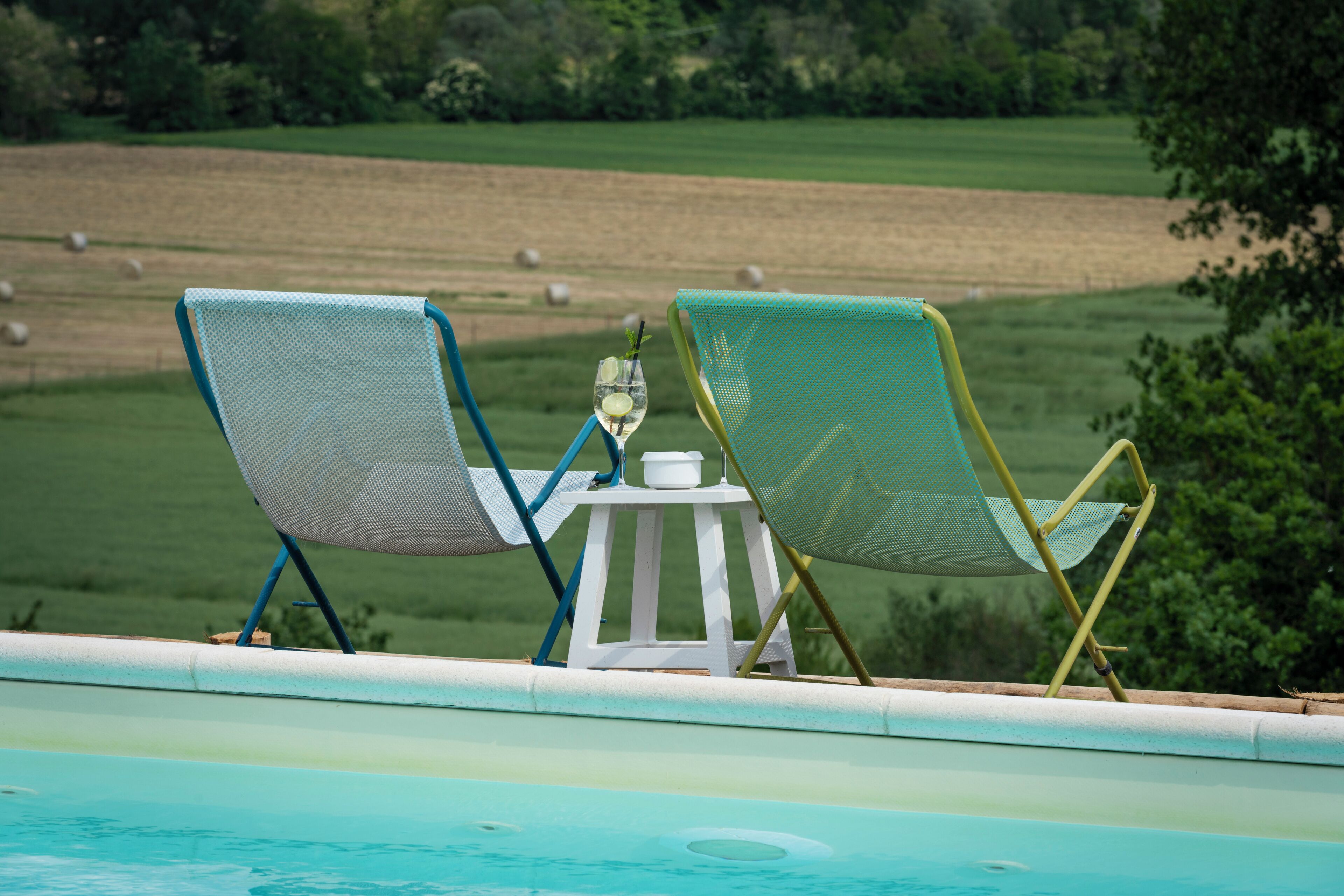 A poolside area with two lounge chairs and a small table with a drink.