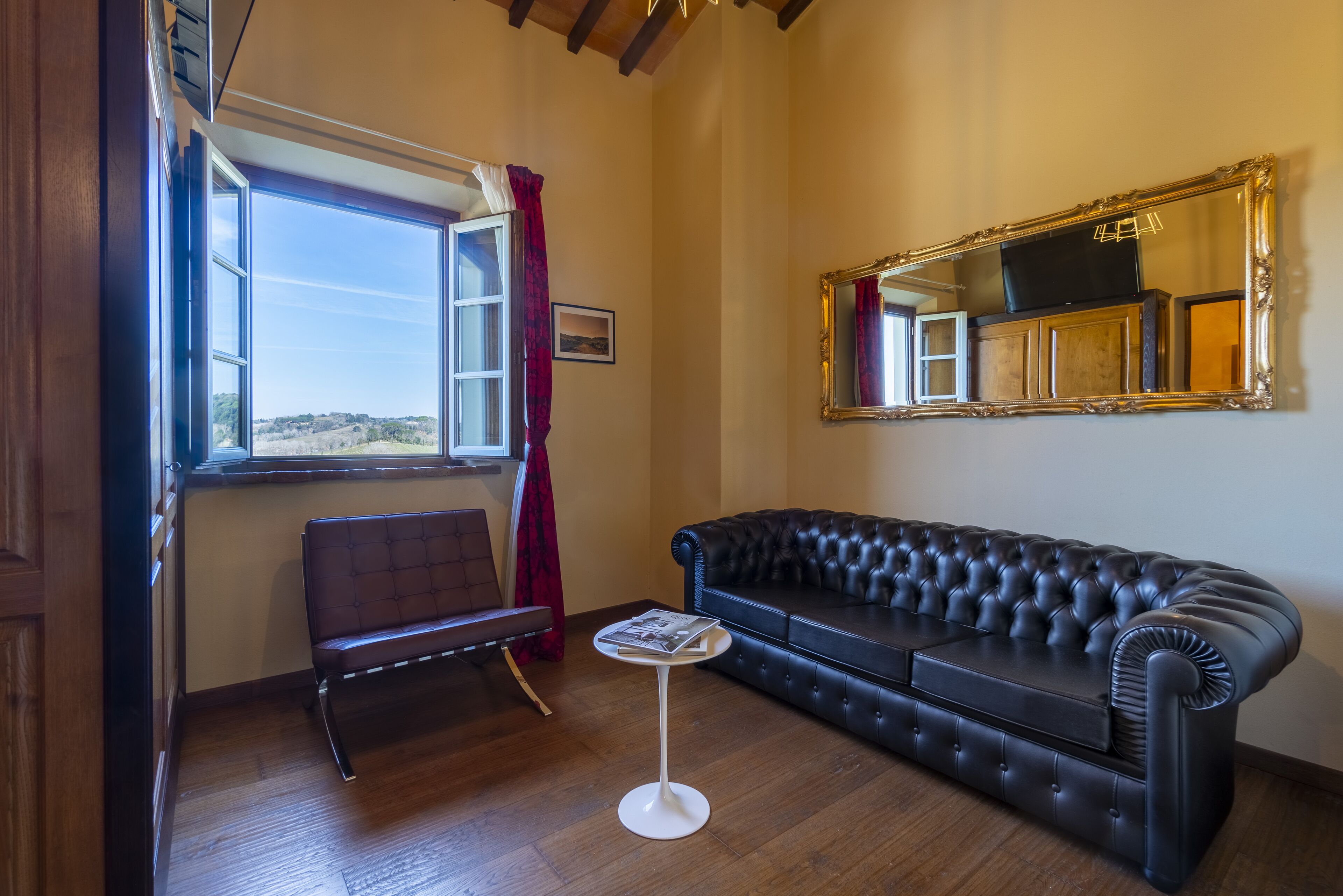 Ein Zimmer mit einem großen Fenster, einem schwarzen Ledersofa, einem kleinen runden Tisch und einem Spiegel an der Wand.