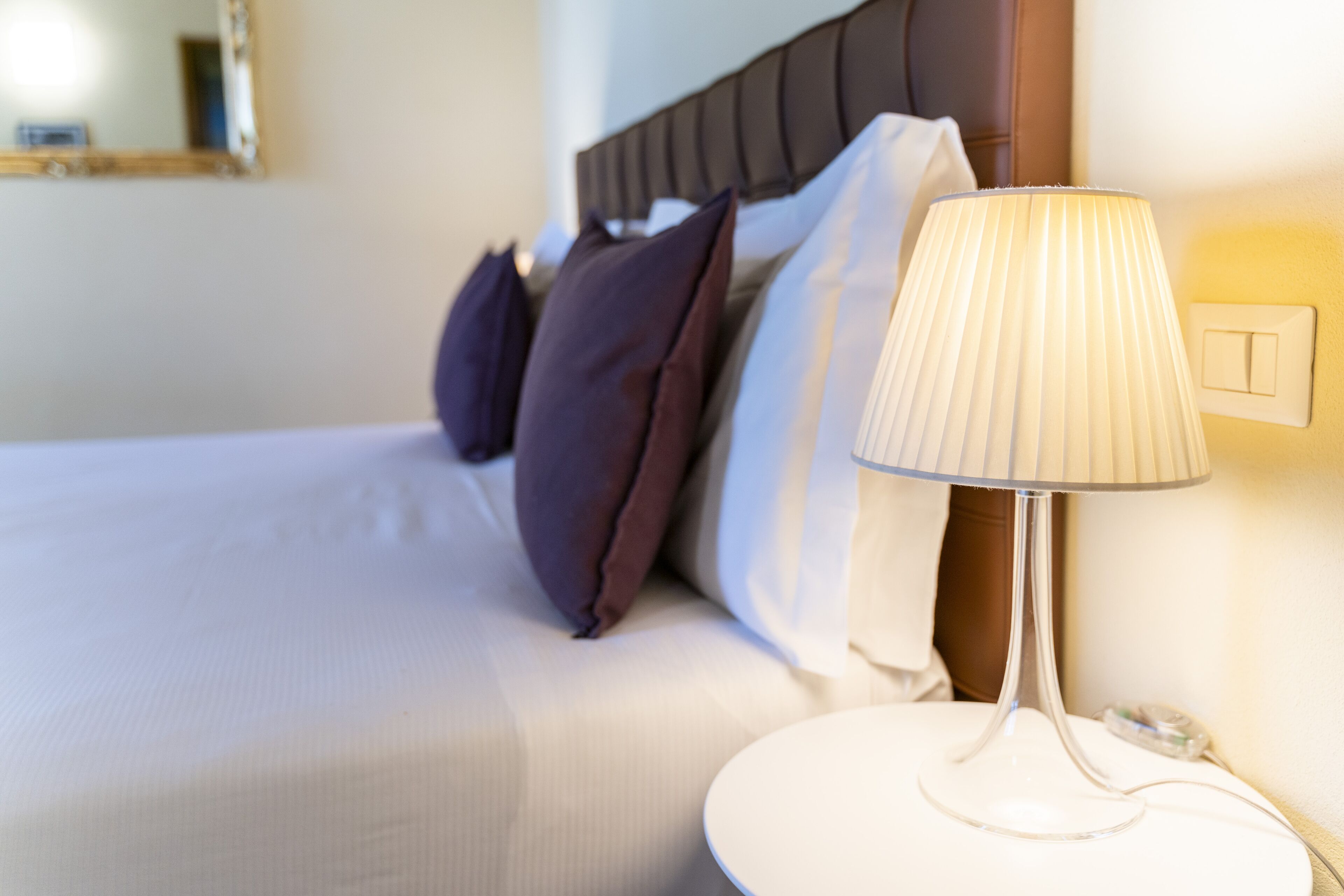 Un lit bien fait, avec une couette blanche et plusieurs oreillers, une table de chevet avec une lampe et un interrupteur fixé au mur.