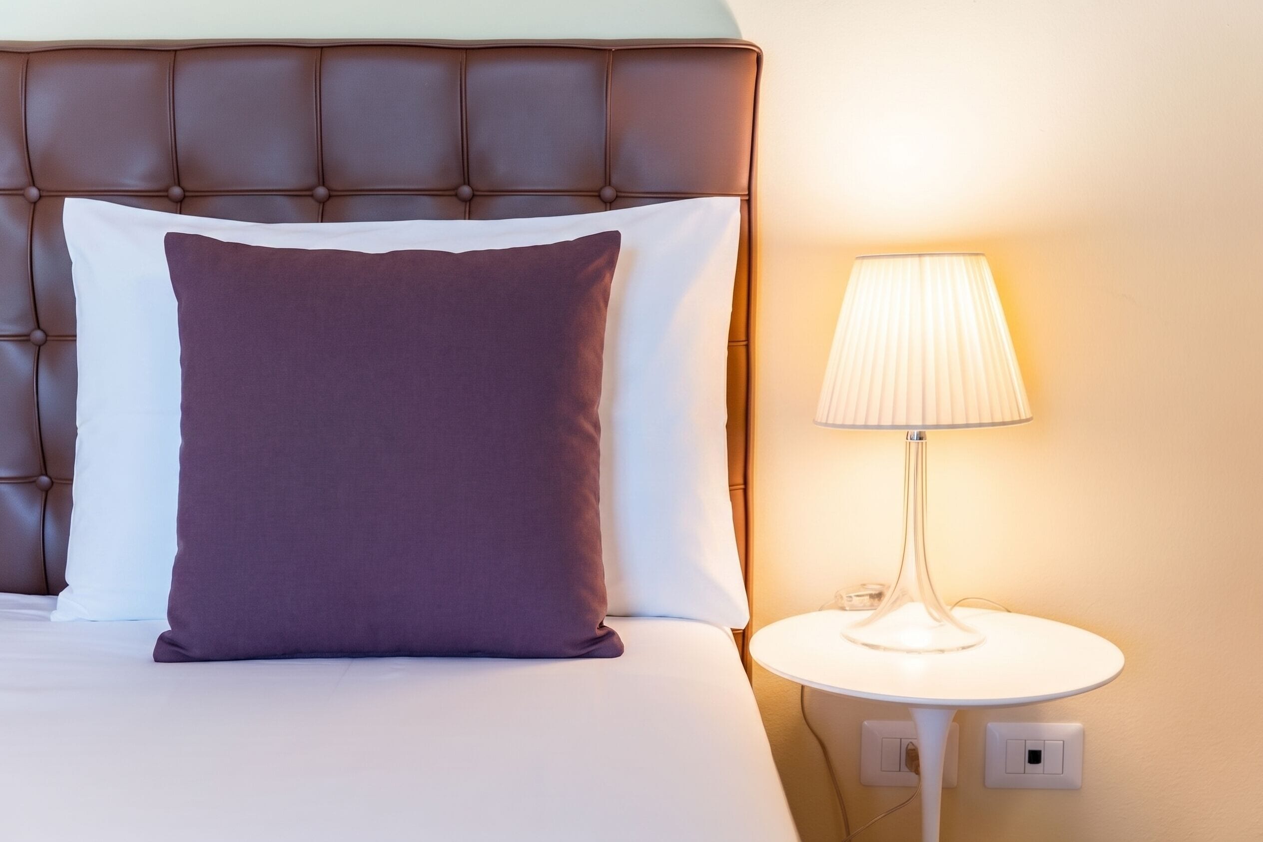 Un lit doté d’une tête de lit capitonnée, d’un oreiller blanc et violet, et d’une lampe de chevet posée sur une table de chevet blanche.