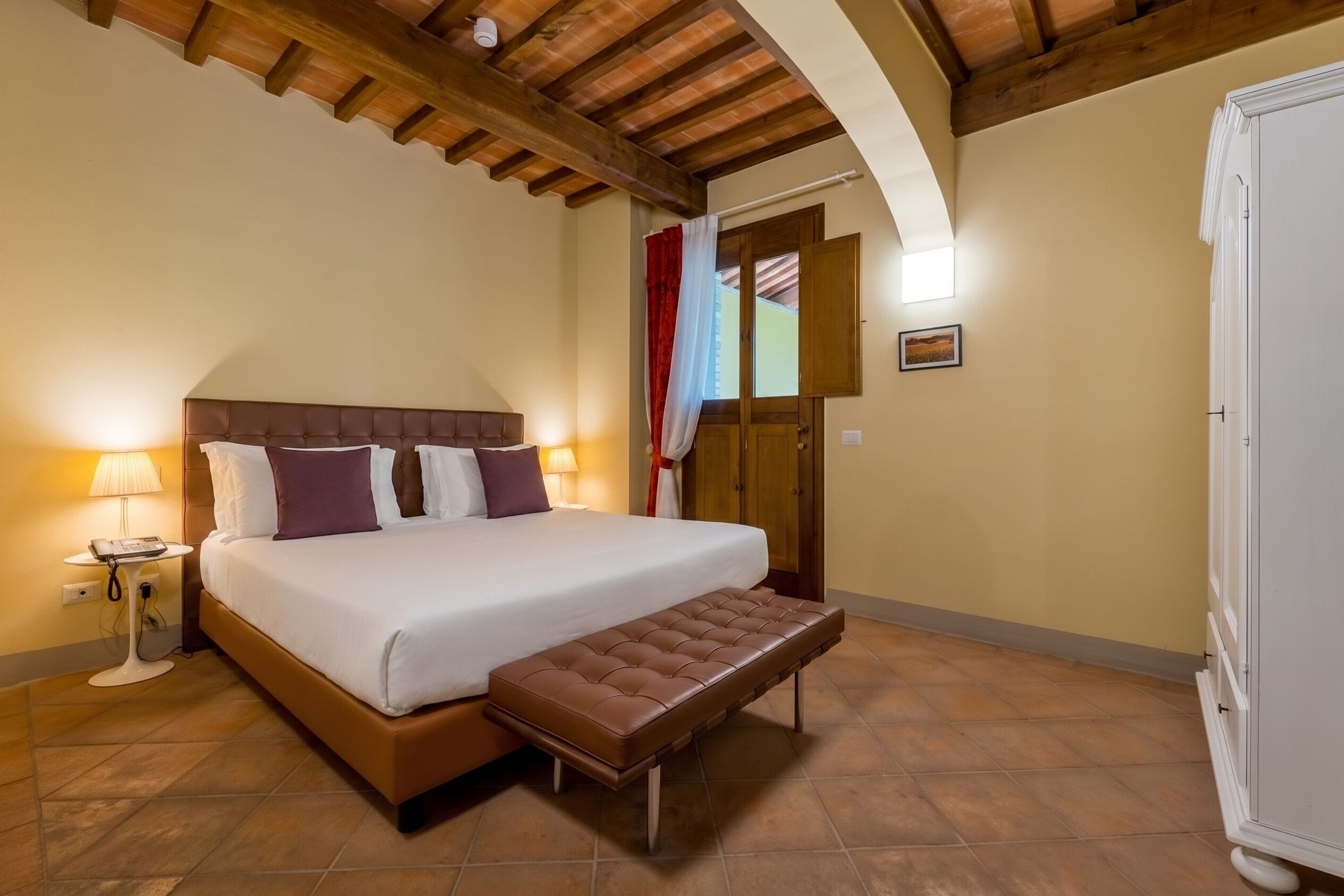 A hotel room with a bed, bedside table, lamp, and a bench.