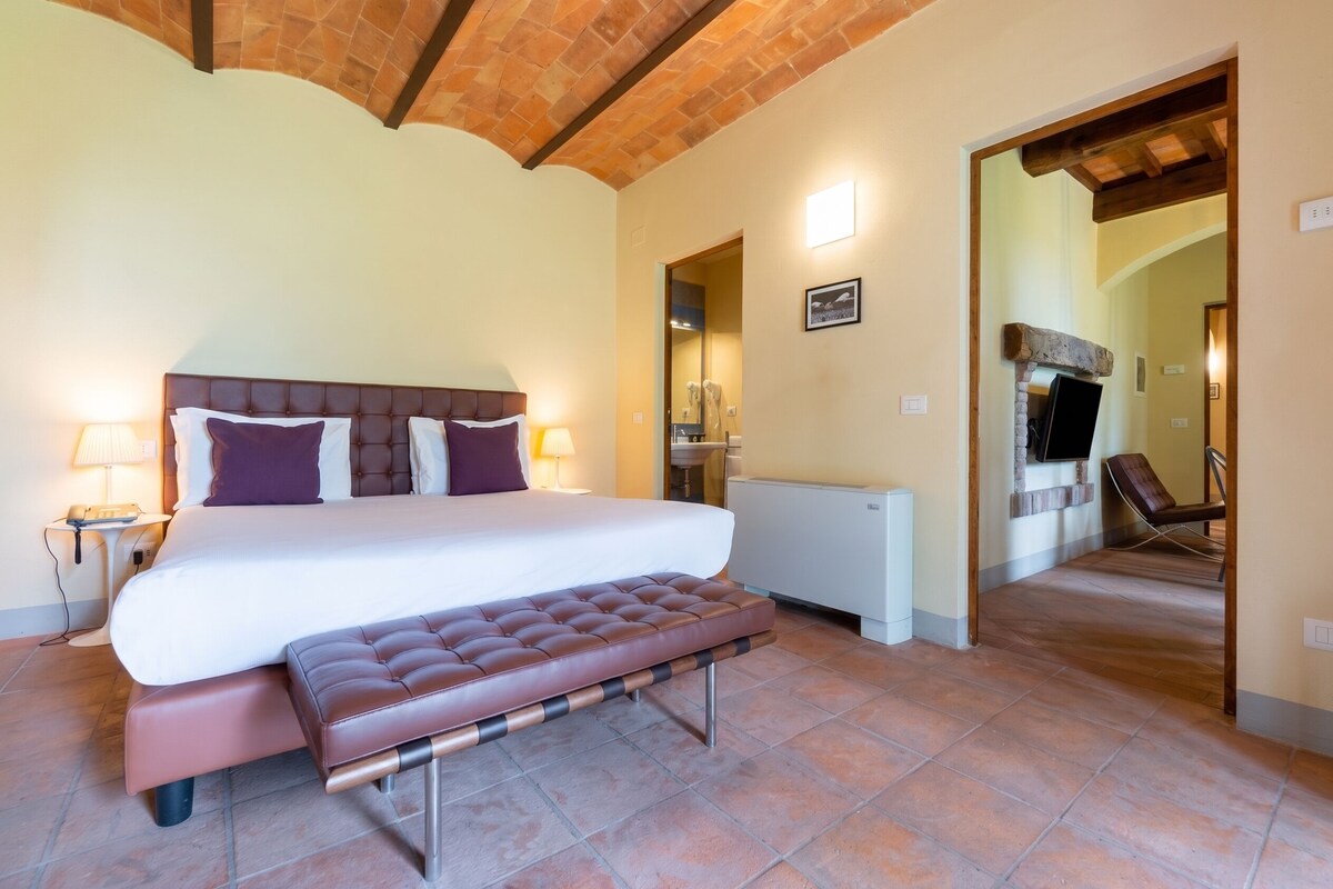 A hotel room with a large bed, a bench, a radiator, a telephone, and a mirror.