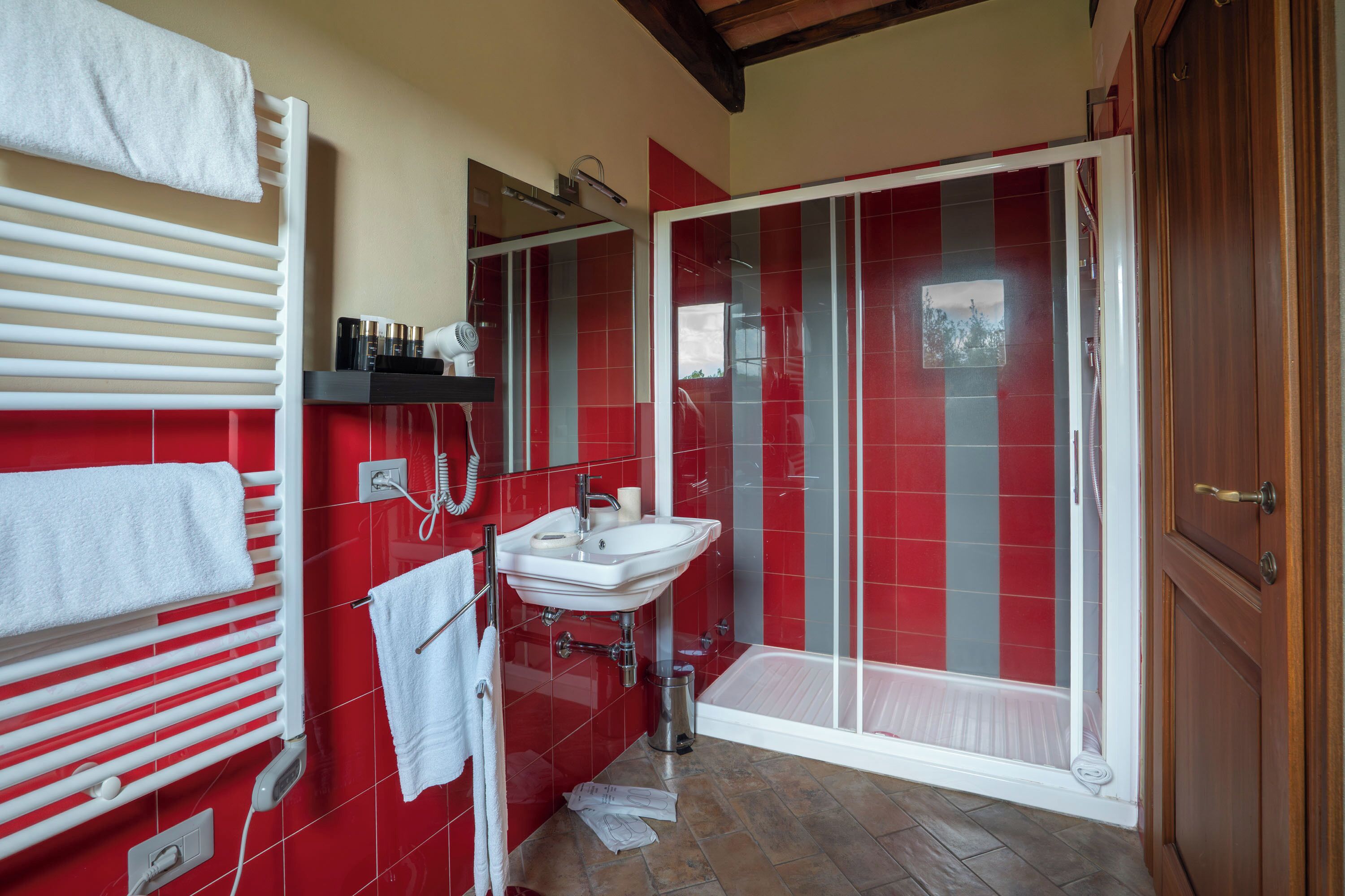 A bathroom with a white sink, red tiled walls, a shower with a glass door, and a towel rack with white towels.