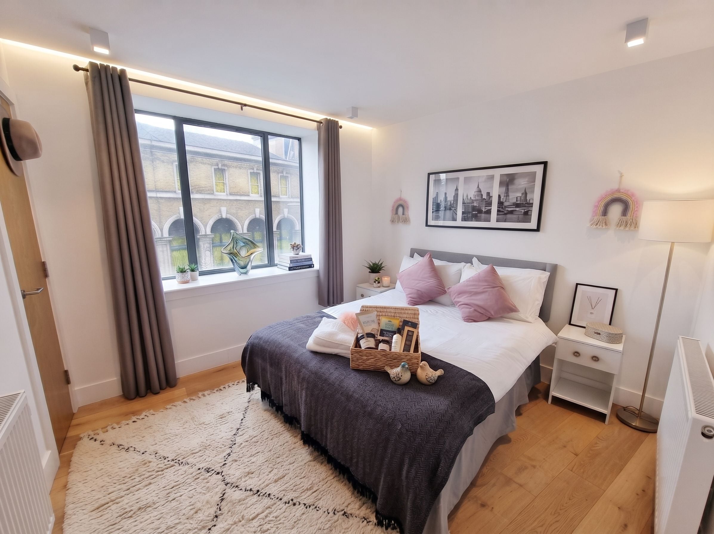 Une chambre avec une grande fenêtre, un lit recouvert d’une couverture grise, une table de chevet avec une lampe et un tableau encadré accroché au mur.