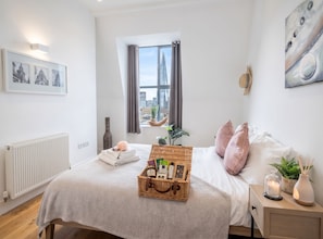 A hotel room with a bed, a nightstand, a window with a city view, a radiator, and a wall with framed pictures. - Urban Stay Shard View Apartments (London)