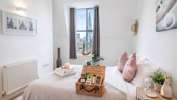 A bedroom with a large window, a bed with pink pillows, a bedside table with a lamp, and a wicker basket on the bed.