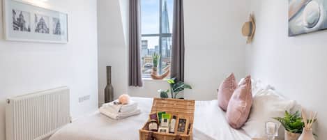 A bedroom with a large window, a bed with pink pillows, a bedside table with a lamp, and a wicker basket on the bed.