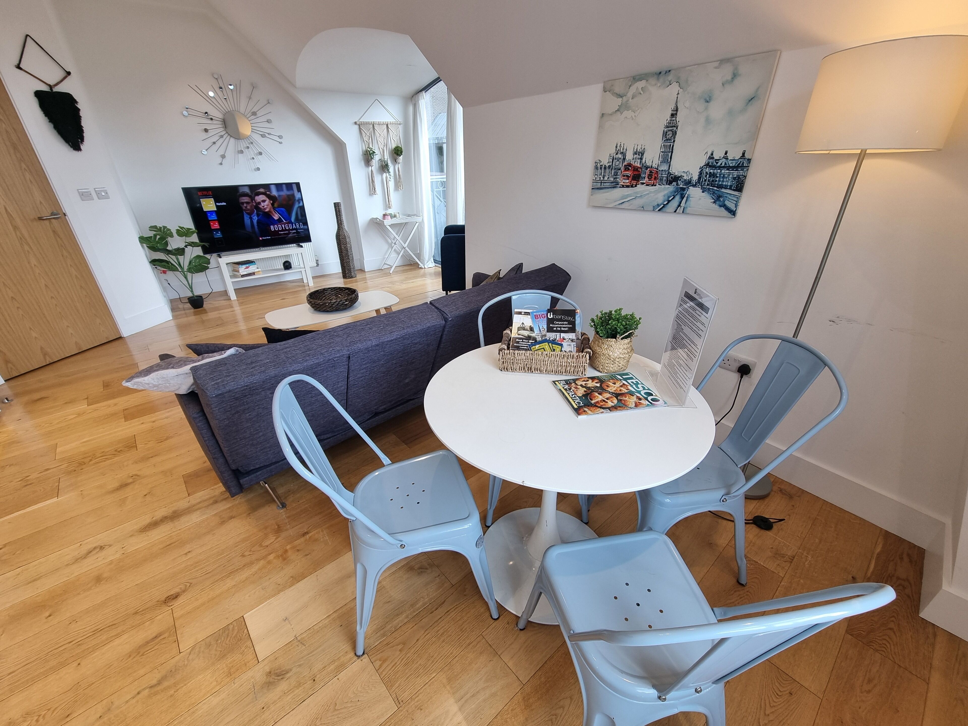 A modern living room with a grey sofa, a round white table, and two blue chairs. There's a TV on the left, a painting of a cityscape on the wall, and a lamp on the right.