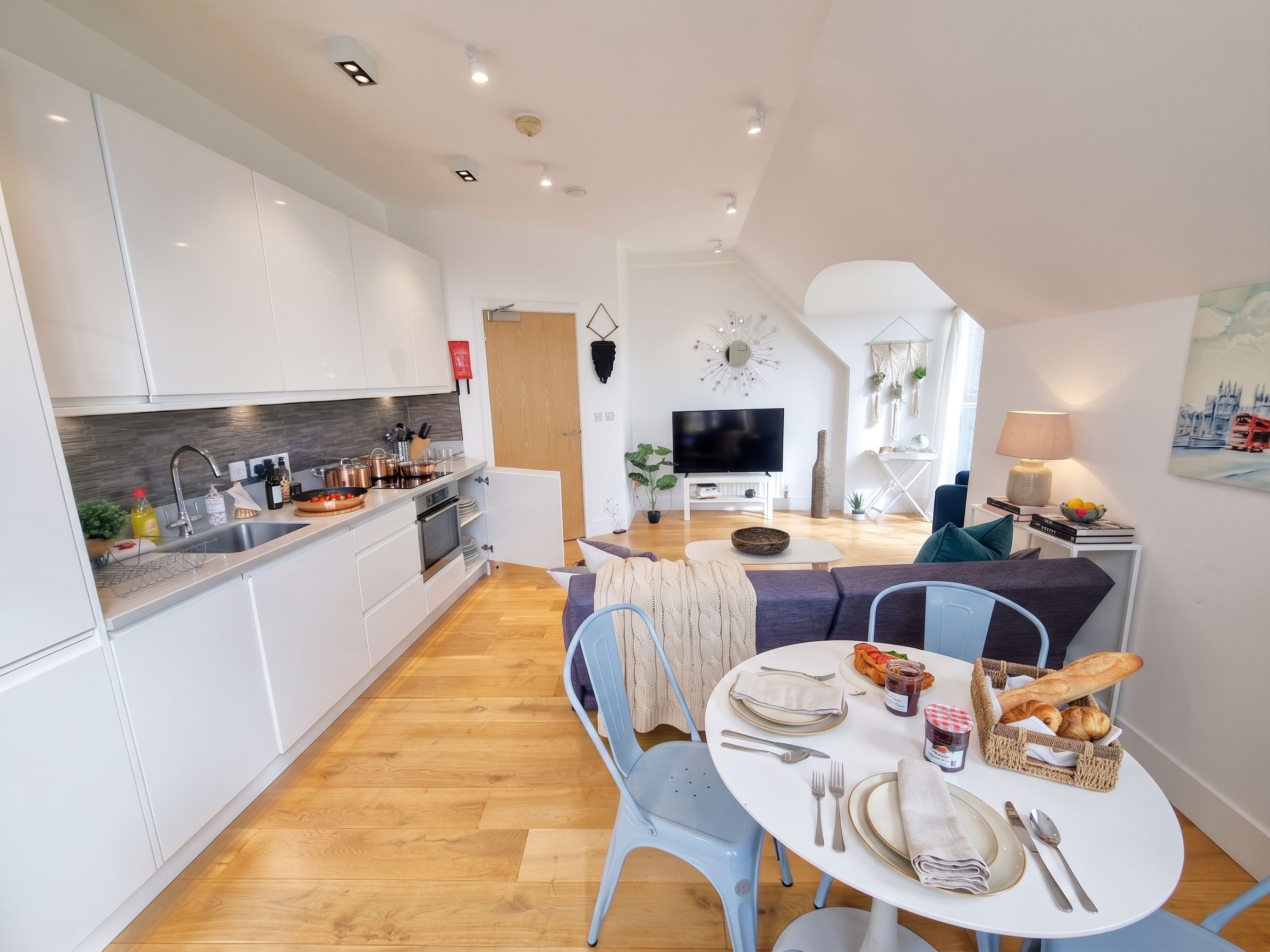Una cocina moderna con armarios blancos, un comedor con mesa redonda y una sala de estar con televisor y asientos.