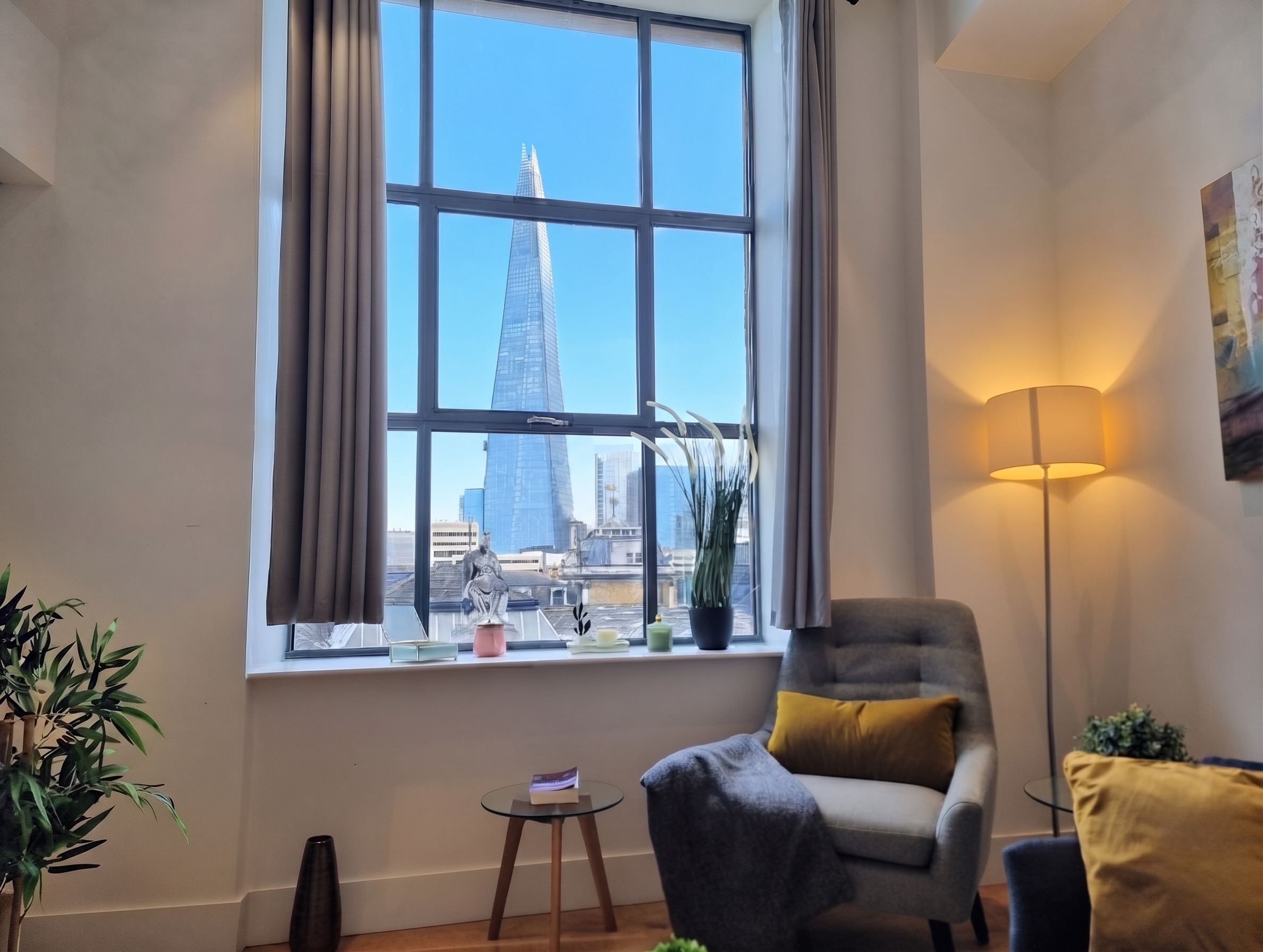 A room with a large window, a chair, a floor lamp, and a small table with a book.