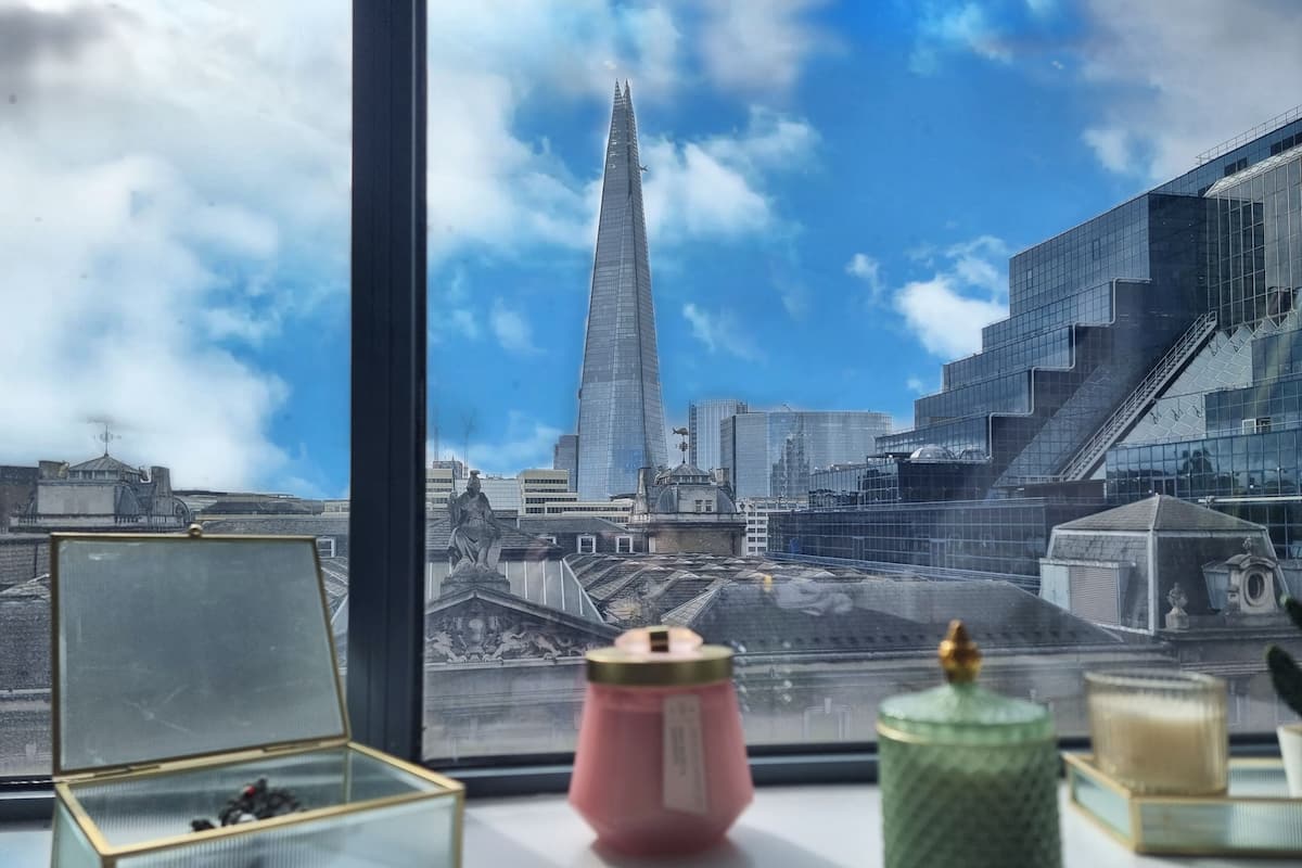 Une vue par la fenêtre sur un paysage urbain moderne, avec un gratte-ciel imposant, un ciel dégagé et une table ornée d’objets décoratifs.