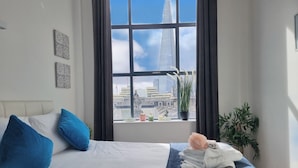A hotel room with a large window, a bed with blue and white bedding, a potted plant, and a view of a tall building. - Urban Stay Shard View Apartments (London)