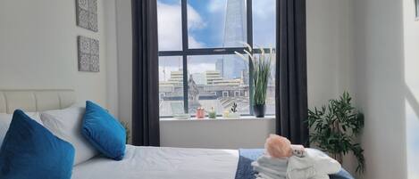 A hotel room with a large window, a bed with blue and white bedding, a potted plant, and a view of a tall building.