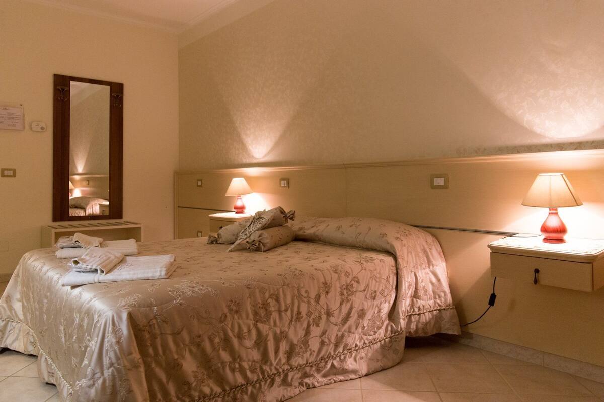 A hotel room with a bed, two bedside lamps, a nightstand, a mirror, and a wall-mounted thermostat.