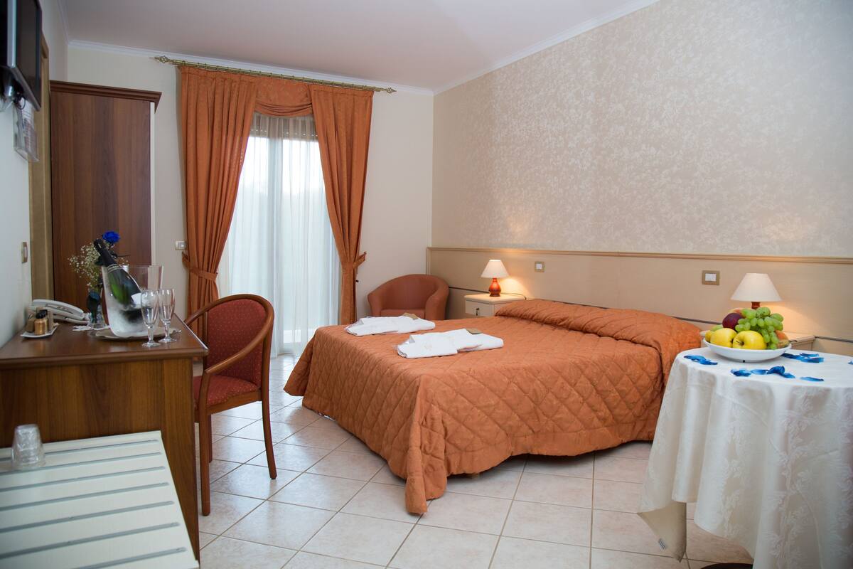 A hotel room with a bed, a chair, a table with fruit, and a lamp.