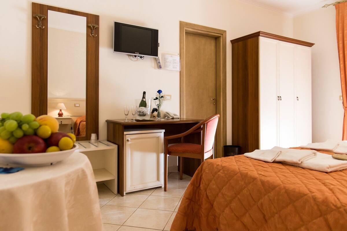 A hotel room with a bed, a desk with a chair, a television, a small refrigerator, and a fruit bowl.
