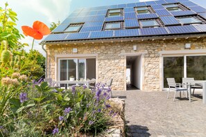 Una casa de piedra con techo de paneles solares, un patio con mobiliario exterior y un jardín con flores moradas.