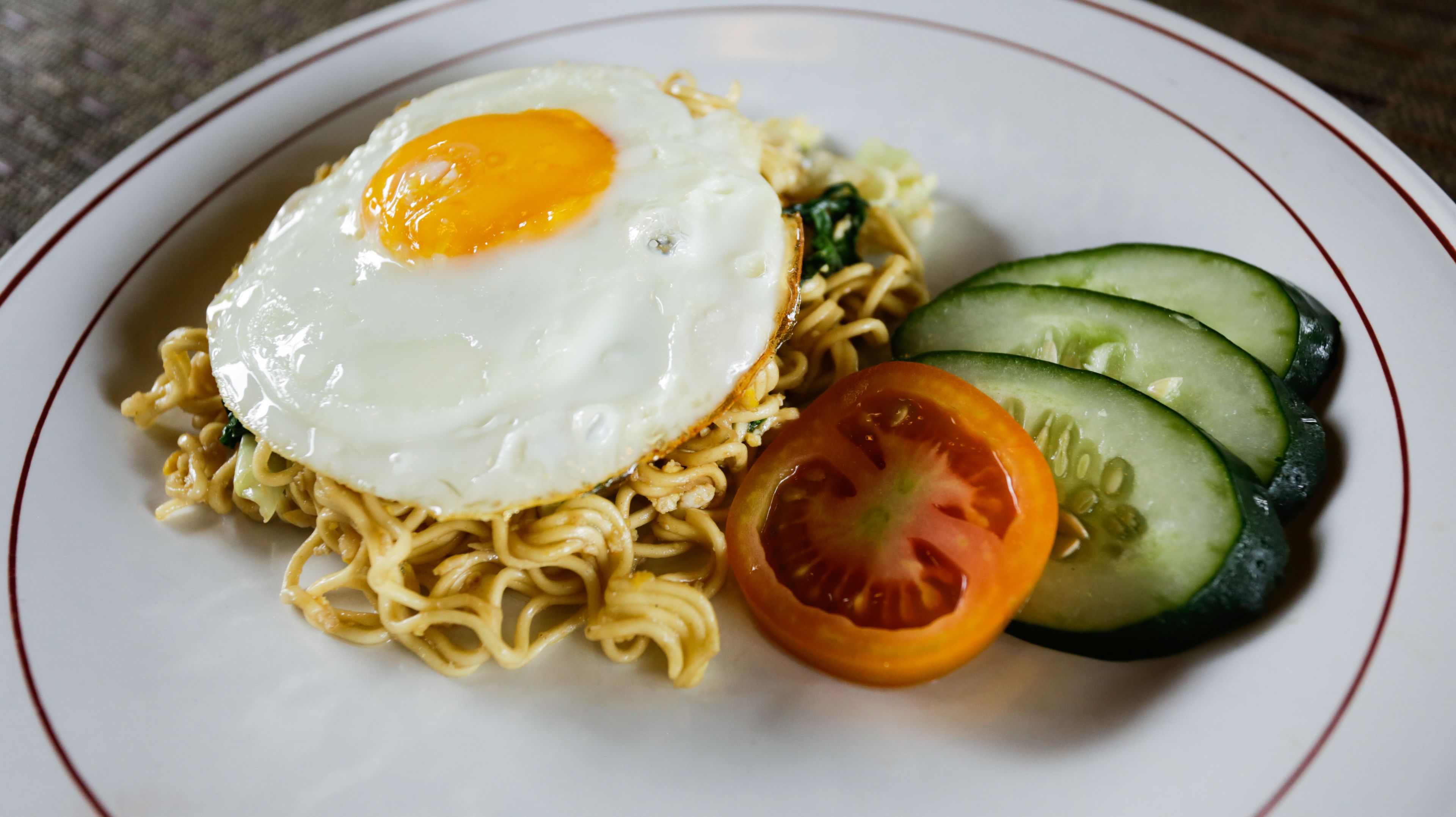 A plate of noodles topped with a fried egg, accompanied by slices of cucumber and tomato.